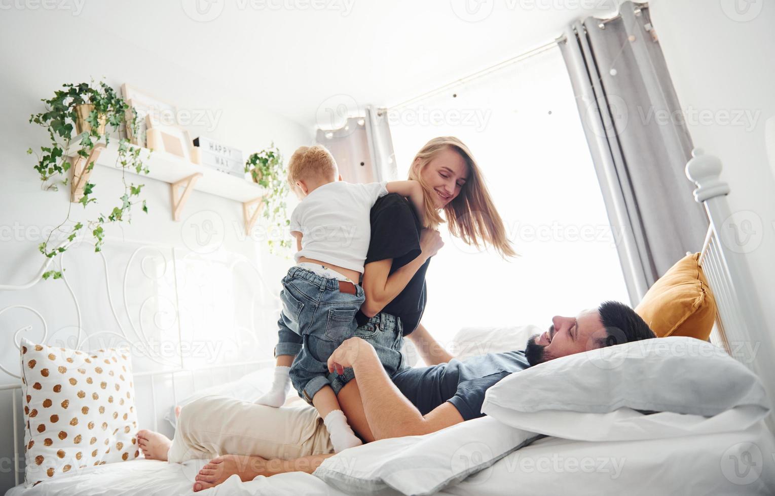 Young married couple with their young son lying down on bed and have fun together in bedroom at daytime photo