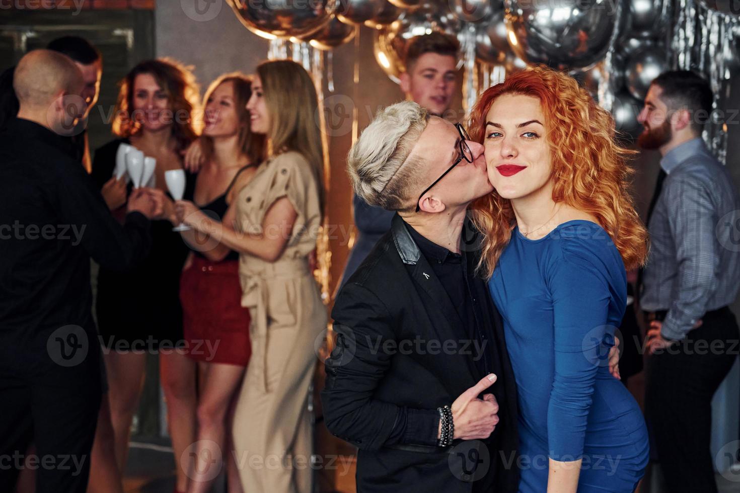 Young man kisses redhead woman against their friends in christmas decorated room and celebrating New Year photo