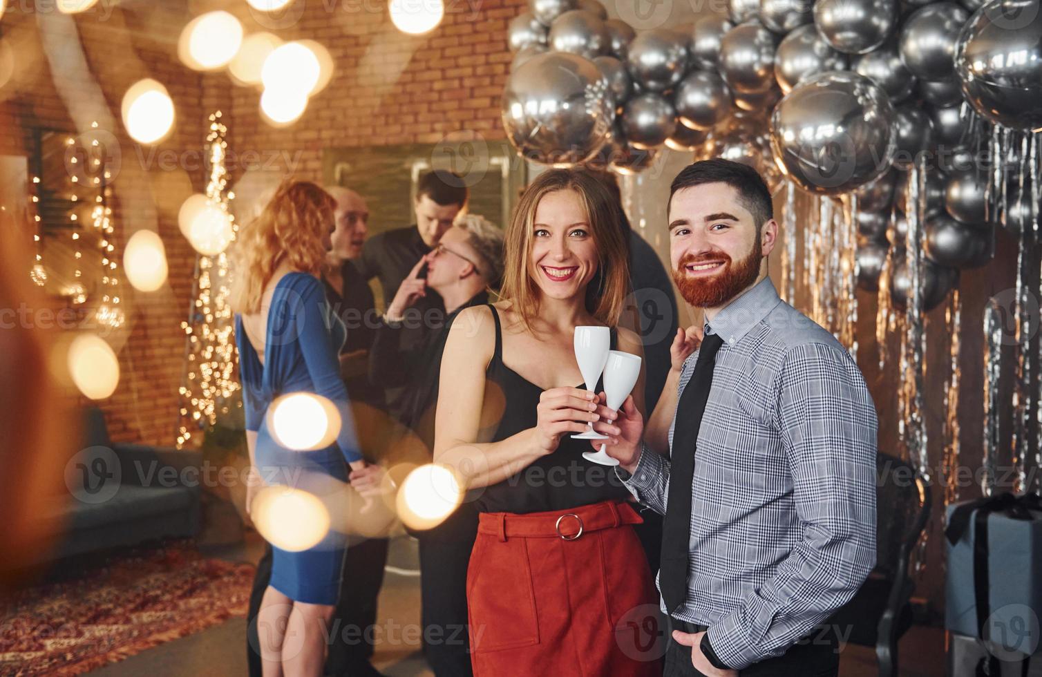 Bearded young man with his girlfriend standing together against their friends in christmas decorated room and celebrating New Year photo