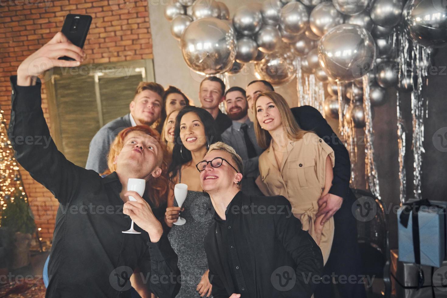 Man taking selfie of his friends that standing in christmas decorated room and celebrating New Year holidays photo