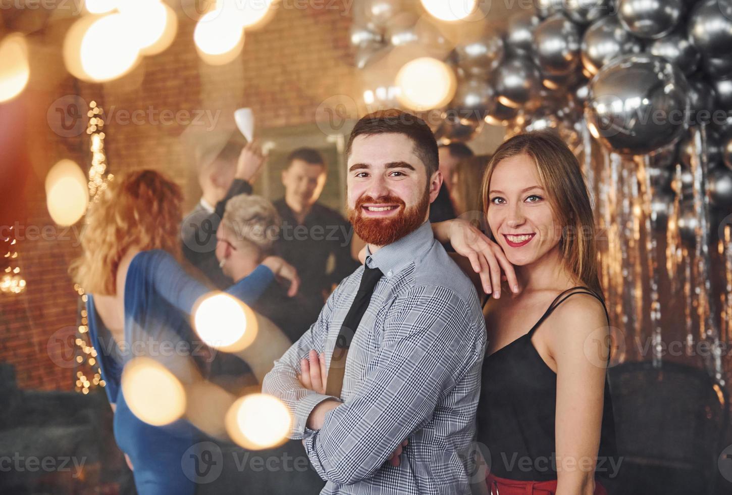 joven barbudo con su novia parados juntos contra sus amigos en una habitación decorada de navidad y celebrando el año nuevo foto