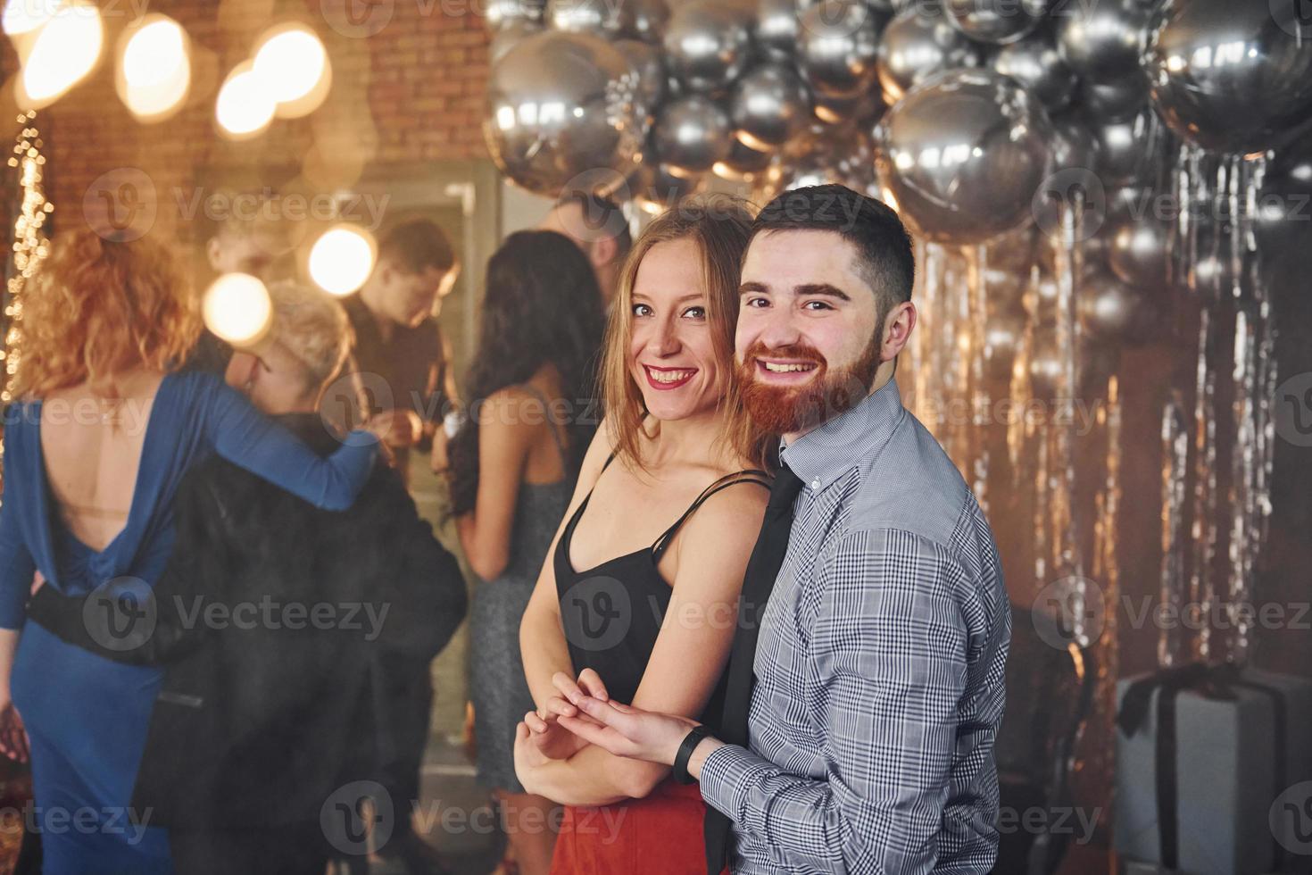 Bearded young man with his girlfriend standing together against their friends in christmas decorated room and celebrating New Year photo