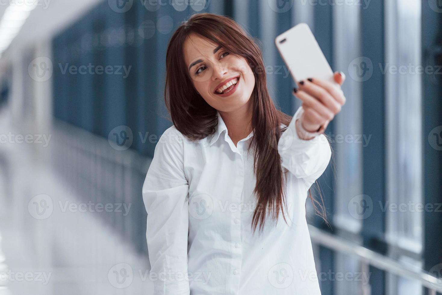 Brunette in white shirt making selfie indoors in modern airport or hallway at daytime photo