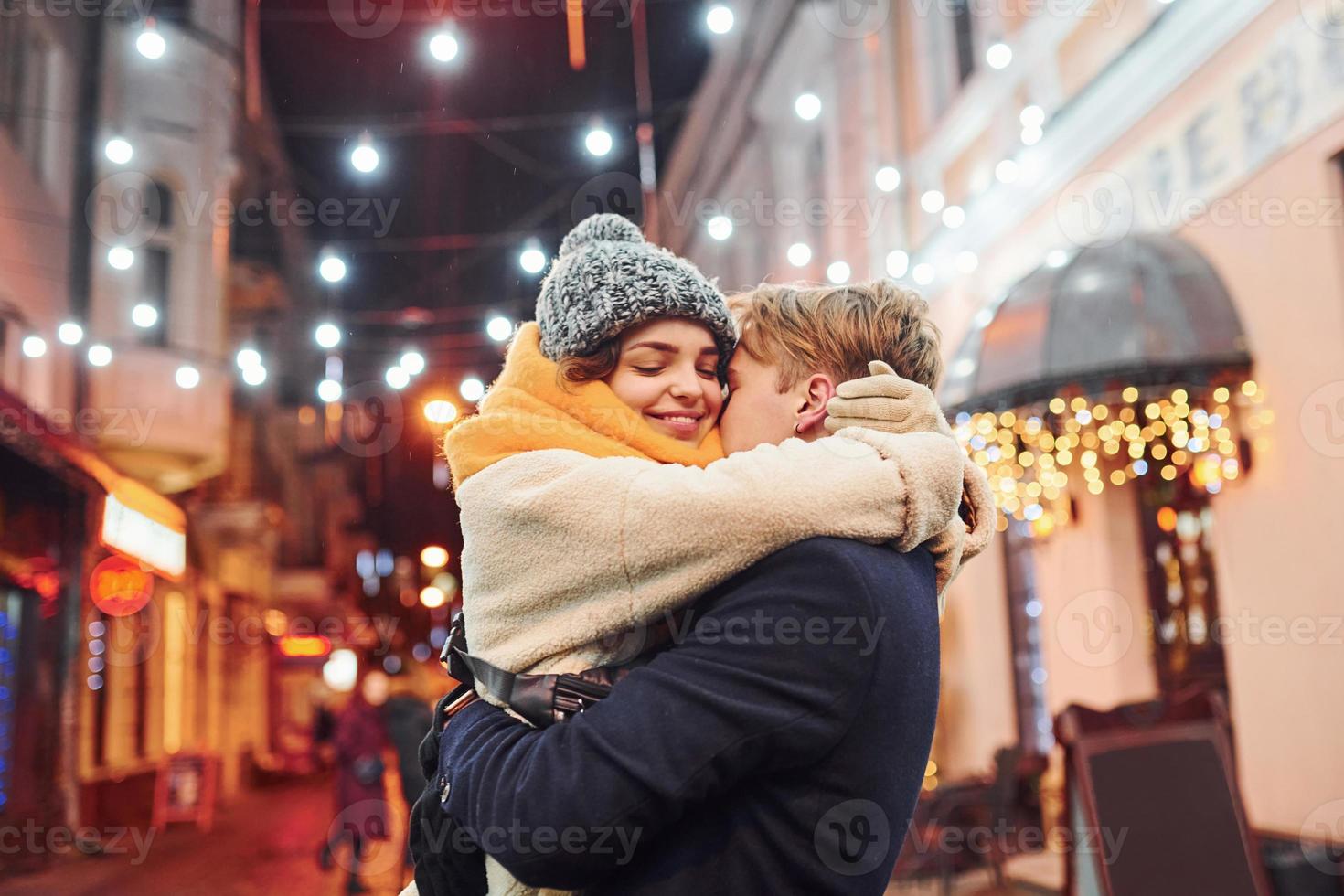 Positive young couple in warm clothes embracing each other on christmas decorated street photo