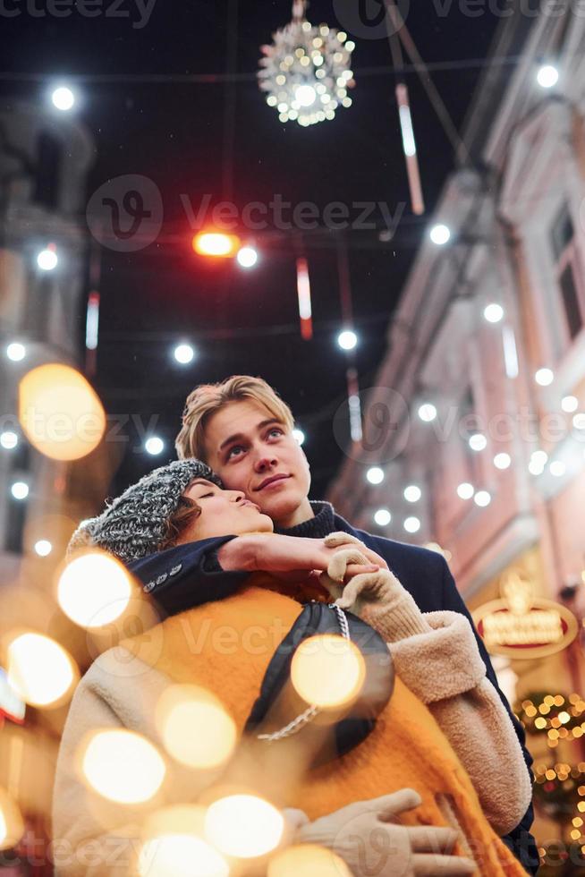 Positive young couple in warm clothes embracing each other on christmas decorated street photo