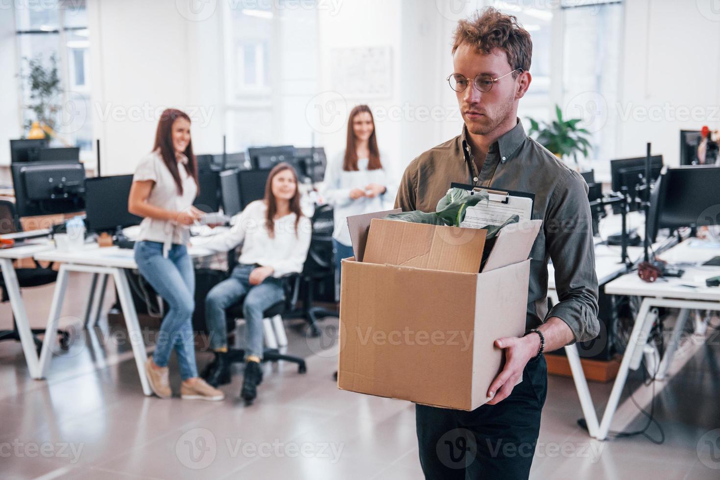 Stylish man in glasses holding box with plant in front of his colleagues in the office photo