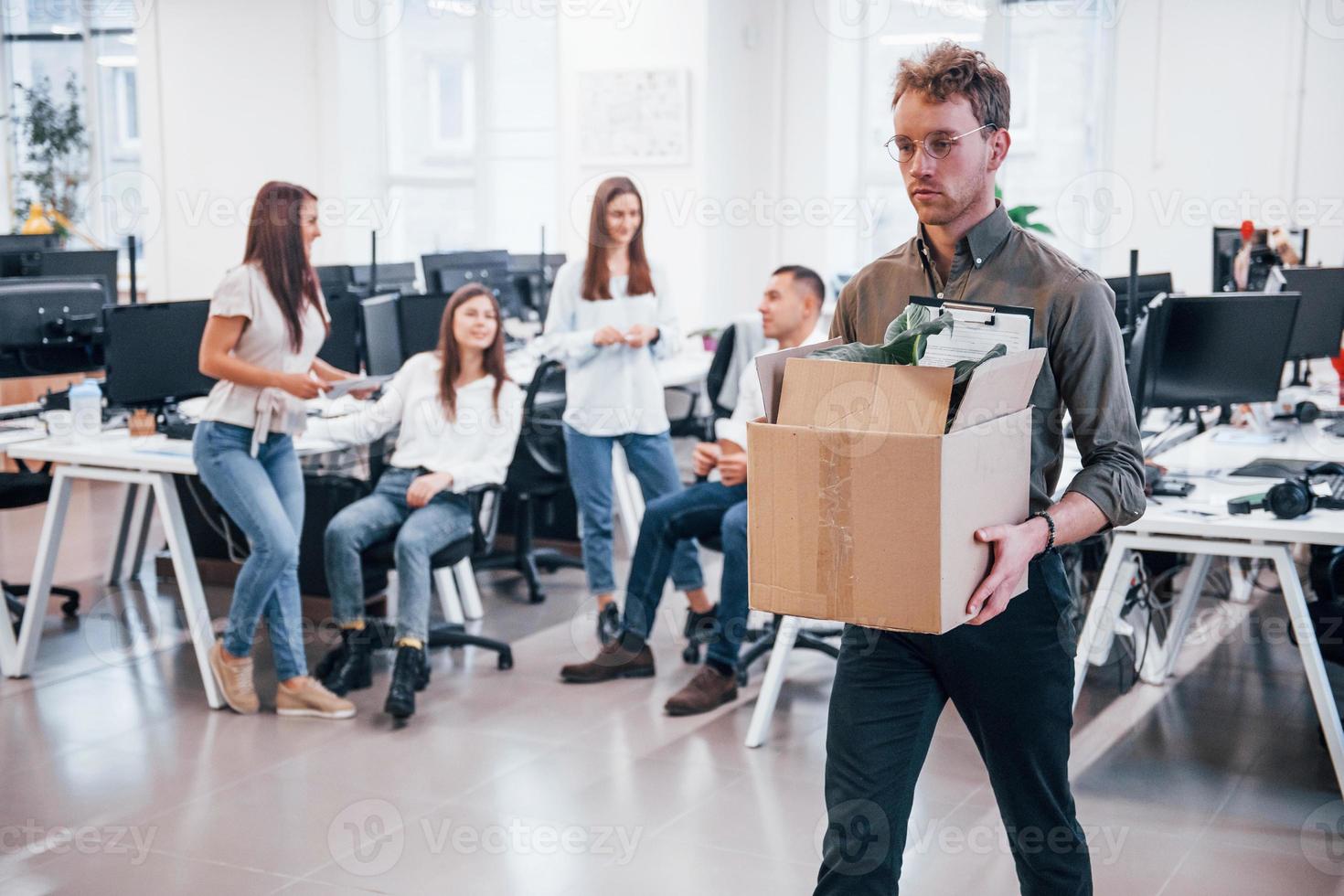 Stylish man in glasses holding box with plant in front of his colleagues in the office photo