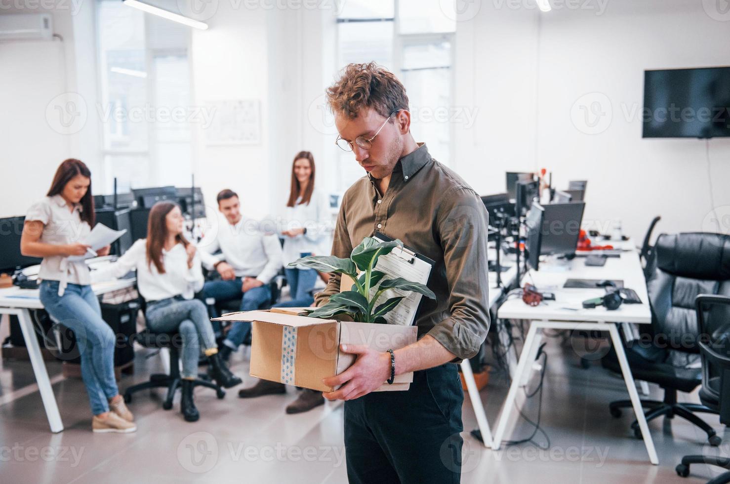Stylish man in glasses holding box with plant in front of his colleagues in the office photo