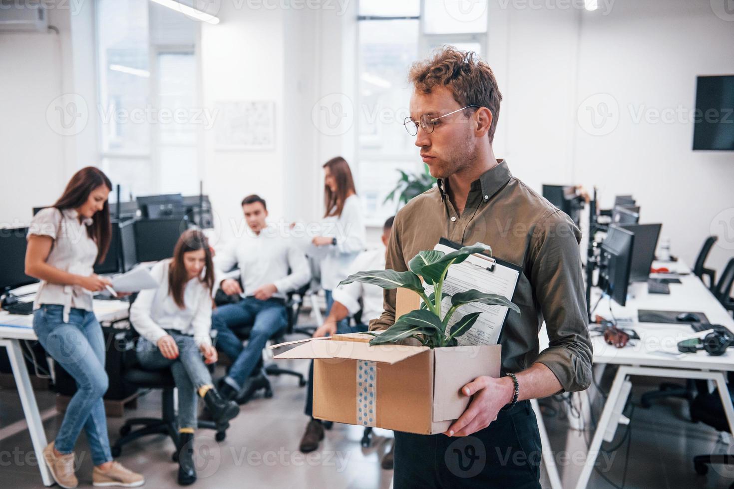Stylish man in glasses holding box with plant in front of his colleagues in the office photo