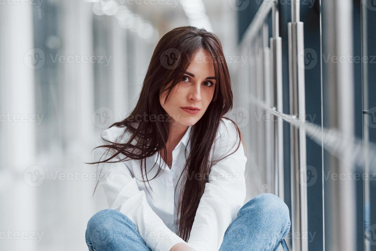 Brunette in white shirt sits indoors in modern airport or hallway at daytime photo