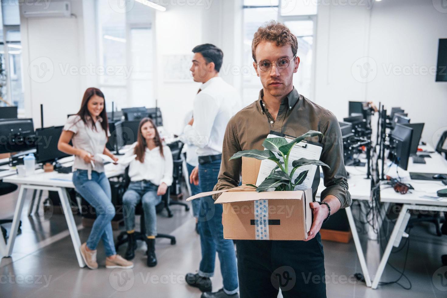 Stylish man in glasses holding box with plant in front of his colleagues in the office photo