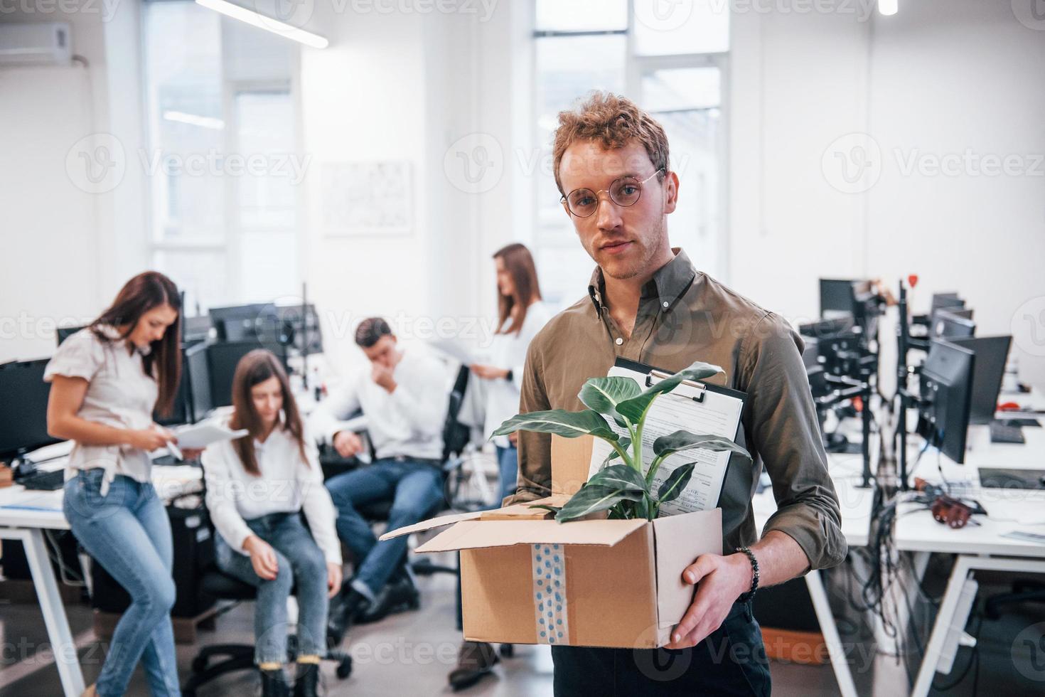 Stylish man in glasses holding box with plant in front of his colleagues in the office photo