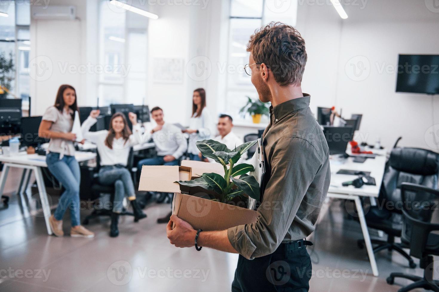 Stylish man in glasses holding box with plant in front of his colleagues in the office photo