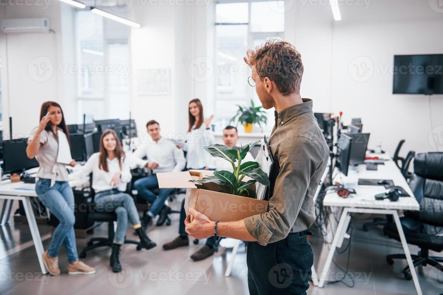 Stylish man in glasses holding box with plant in front of his colleagues in the office photo