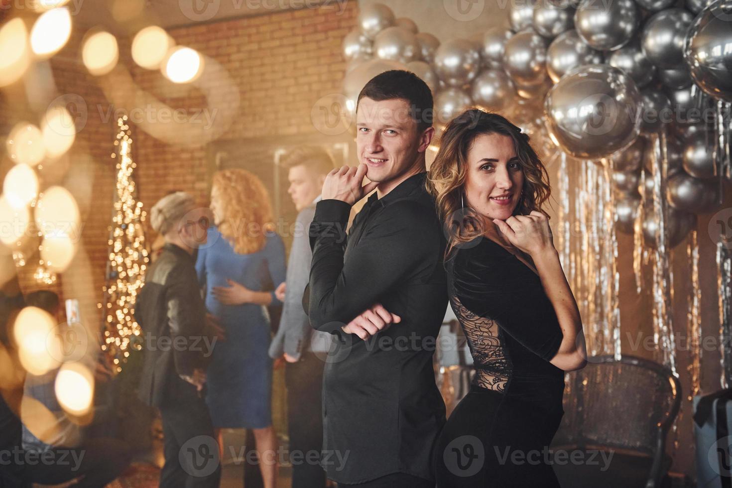 Young couple standing together against their friends in christmas decorated room and celebrating New Year photo