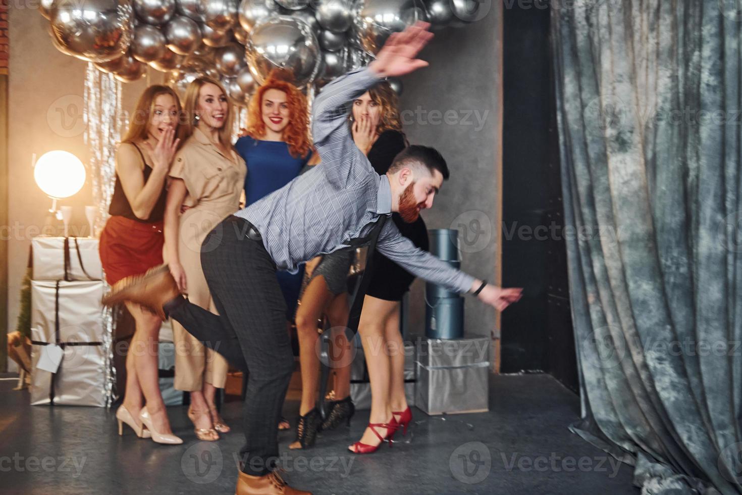 Young women in festive clothes standing indoors of christmas decorated room together and looks at man that have fun doing tricks photo