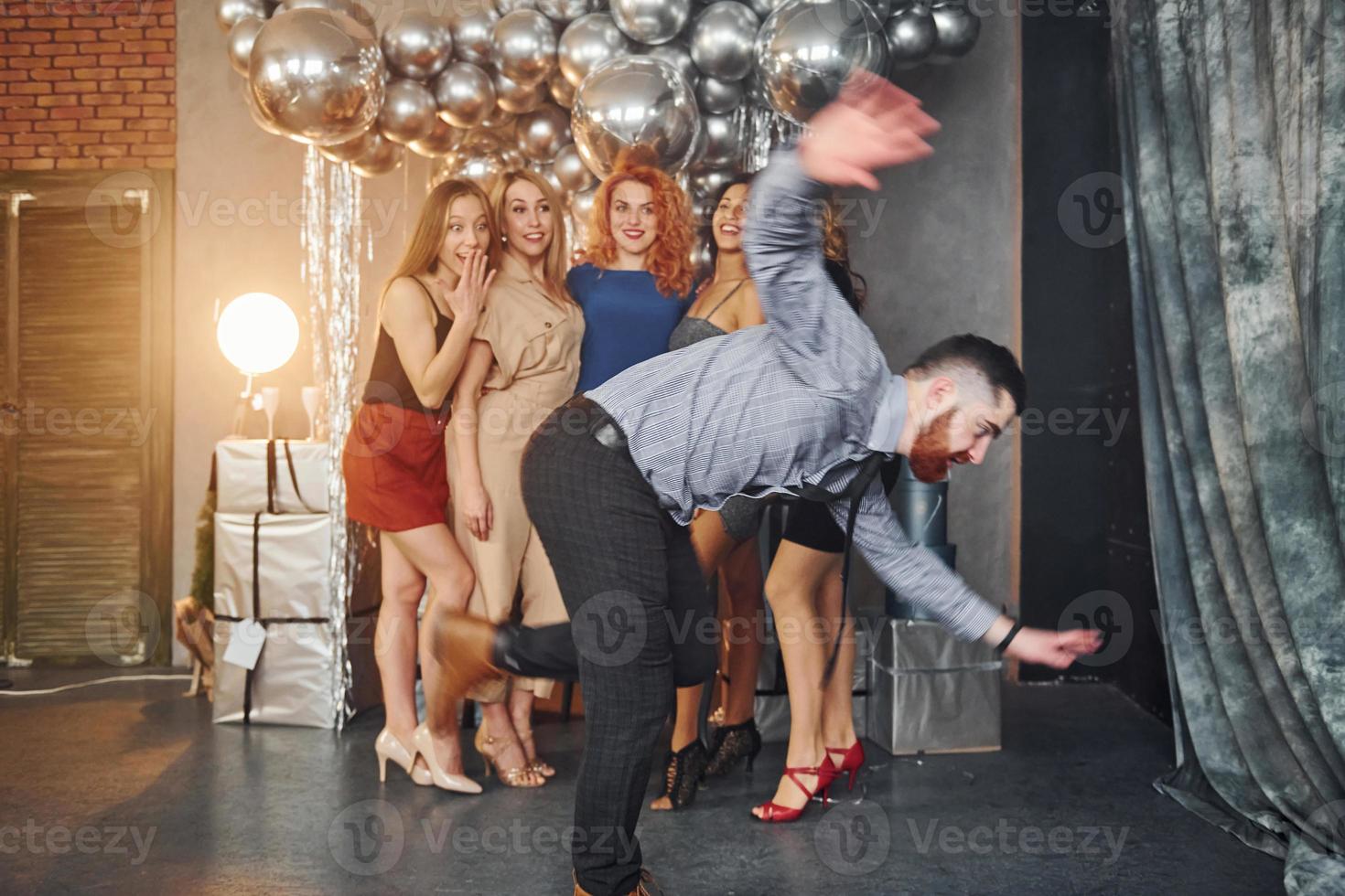 Young women in festive clothes standing indoors of christmas decorated room together and looks at man that have fun doing tricks photo