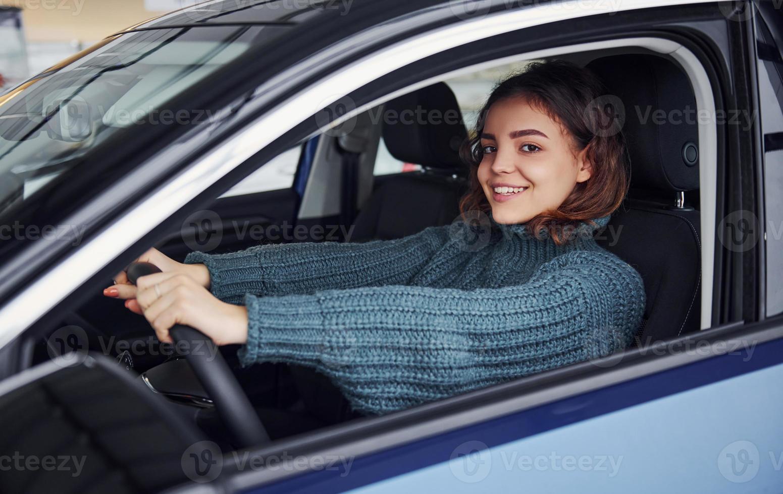 Positive young girl in casual clothes sitting inside of modern car and smiling photo