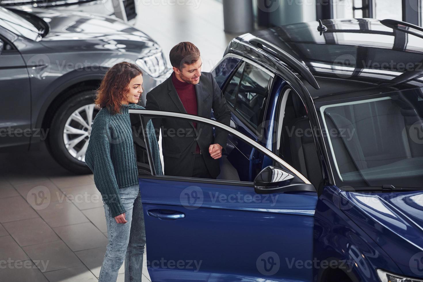Professional salesman assisting young girl by choosing new modern automobile indoors photo