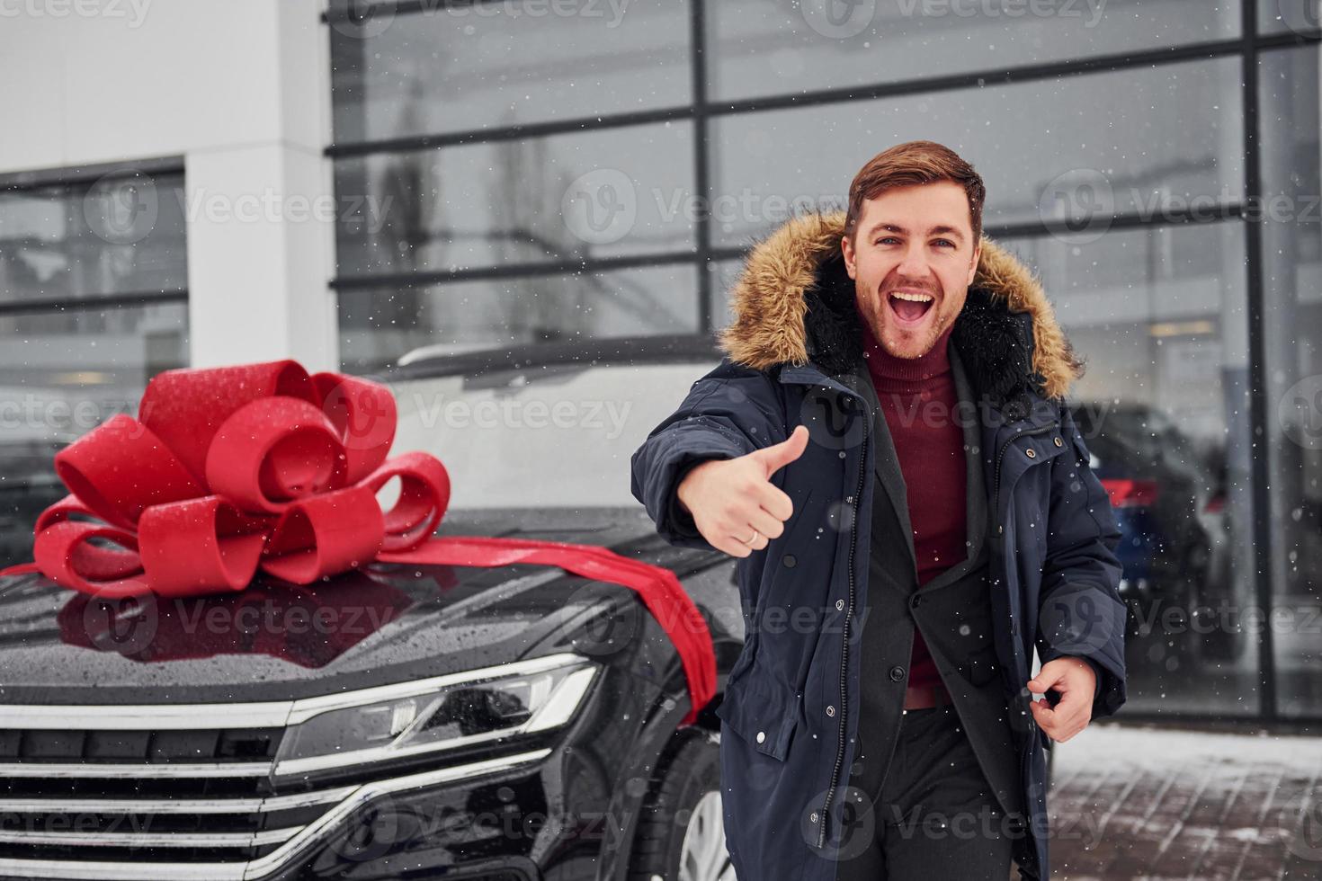 Happy owner of new modern car standing outdoors and smiling. Red gift length on the automobile photo