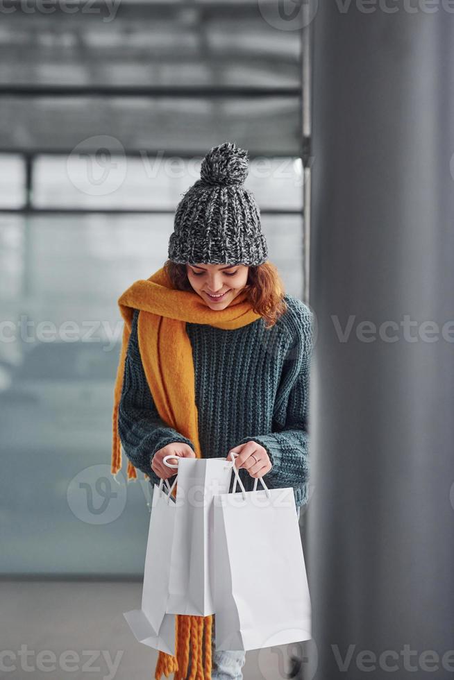 Beautiful cheerful girl in yellow scarf and in warm clothes standing indoors with shopping bags in hands photo