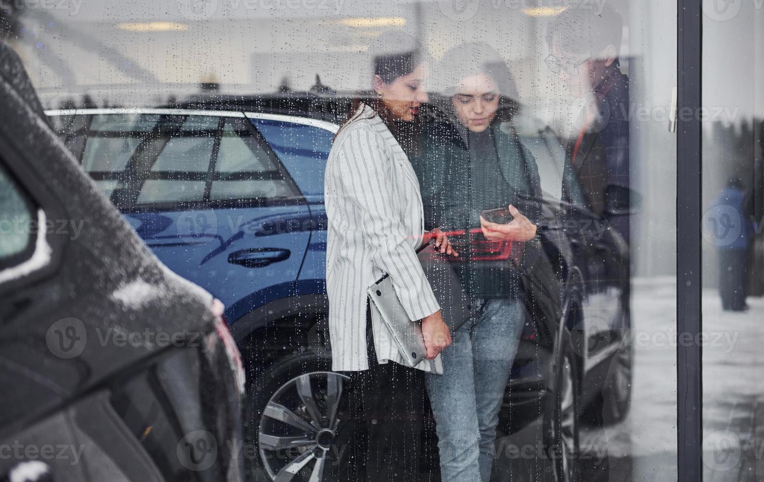 Friends standing behind glass at rainy day. Girl holds laptop in hands photo