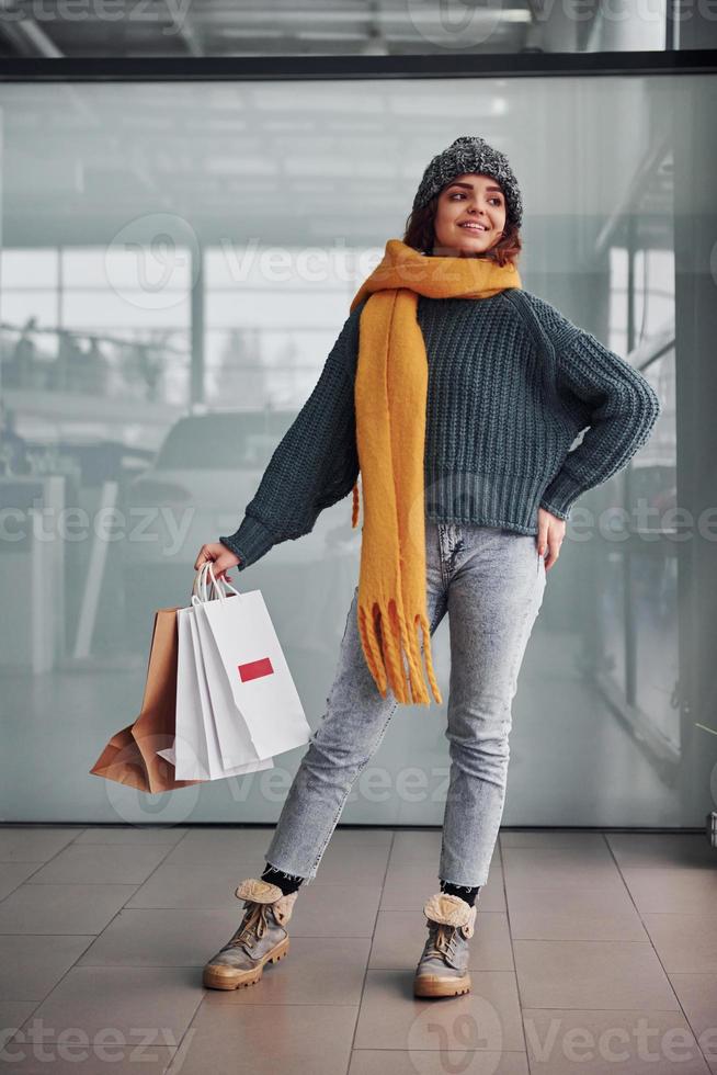 Beautiful cheerful girl in yellow scarf and in warm clothes standing indoors with shopping bags in hands photo