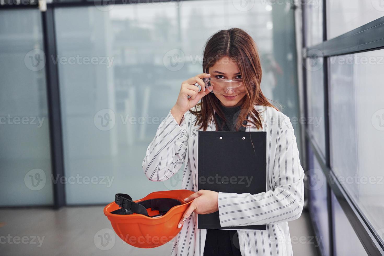 Portrait of young female worker with protective helmet, eyewear and notepad. Standing in the office photo
