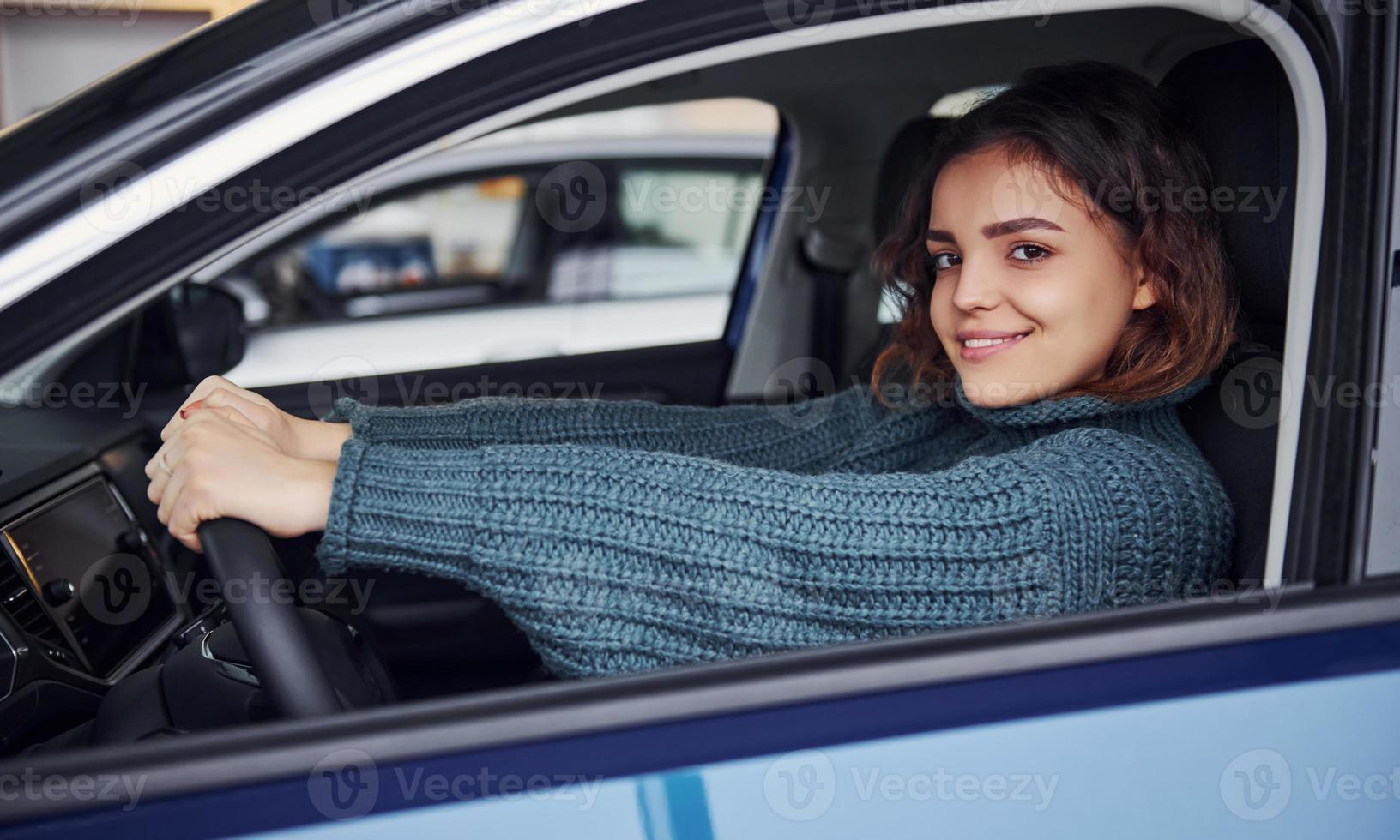 Positive young girl in casual clothes sitting inside of modern car and smiling photo