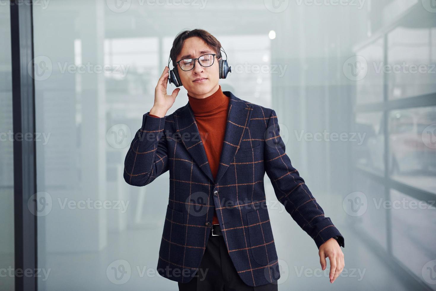 Listening to the music in headphones. Young business man in luxury suit and formal clothes is indoors in the office photo