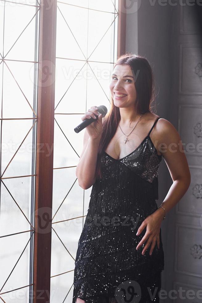 bella mujer con vestido negro de lujo parada cerca de la ventana con micrófono en las manos foto