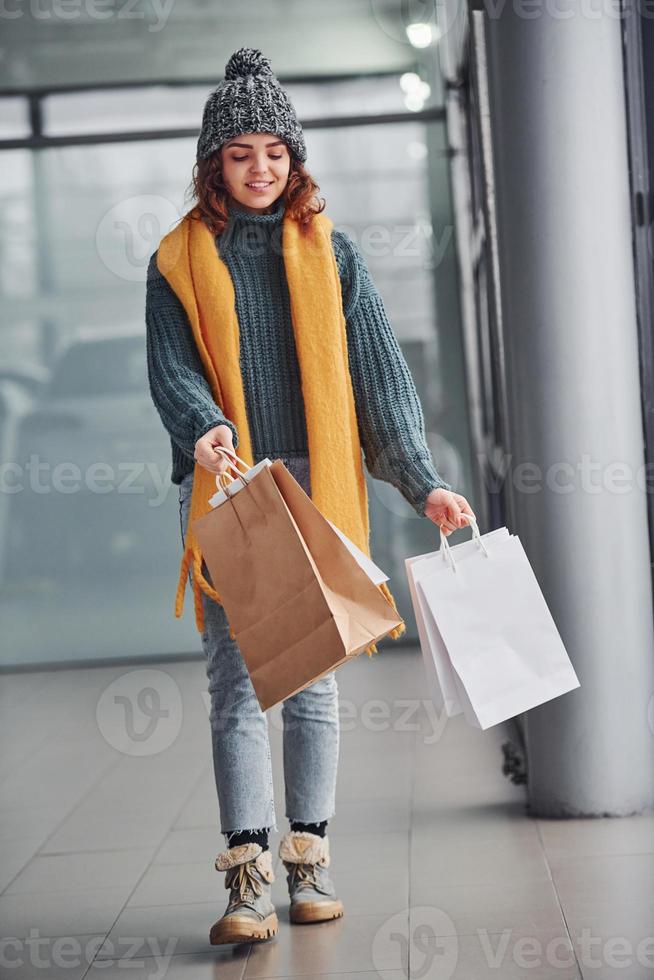 Beautiful cheerful girl in yellow scarf and in warm clothes standing indoors with shopping bags in hands photo