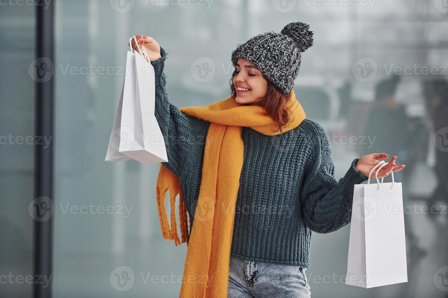 Smiling and feeling happy. Beautiful cheerful girl in yellow scarf and in warm clothes standing indoors with shopping bags in hands photo