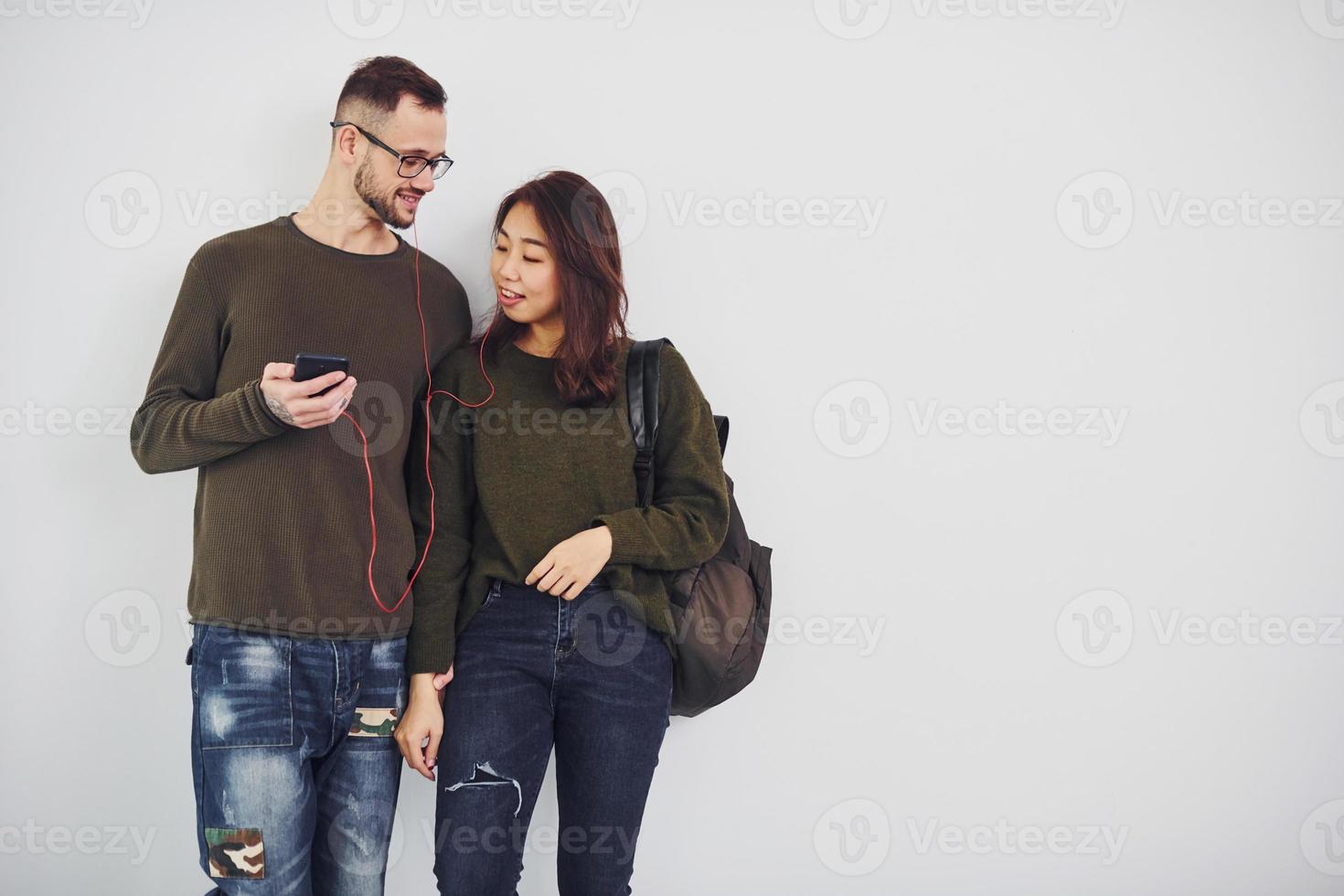 Cheerful multi ethnic couple with backpack and phone standing together indoors in the studio against white background photo