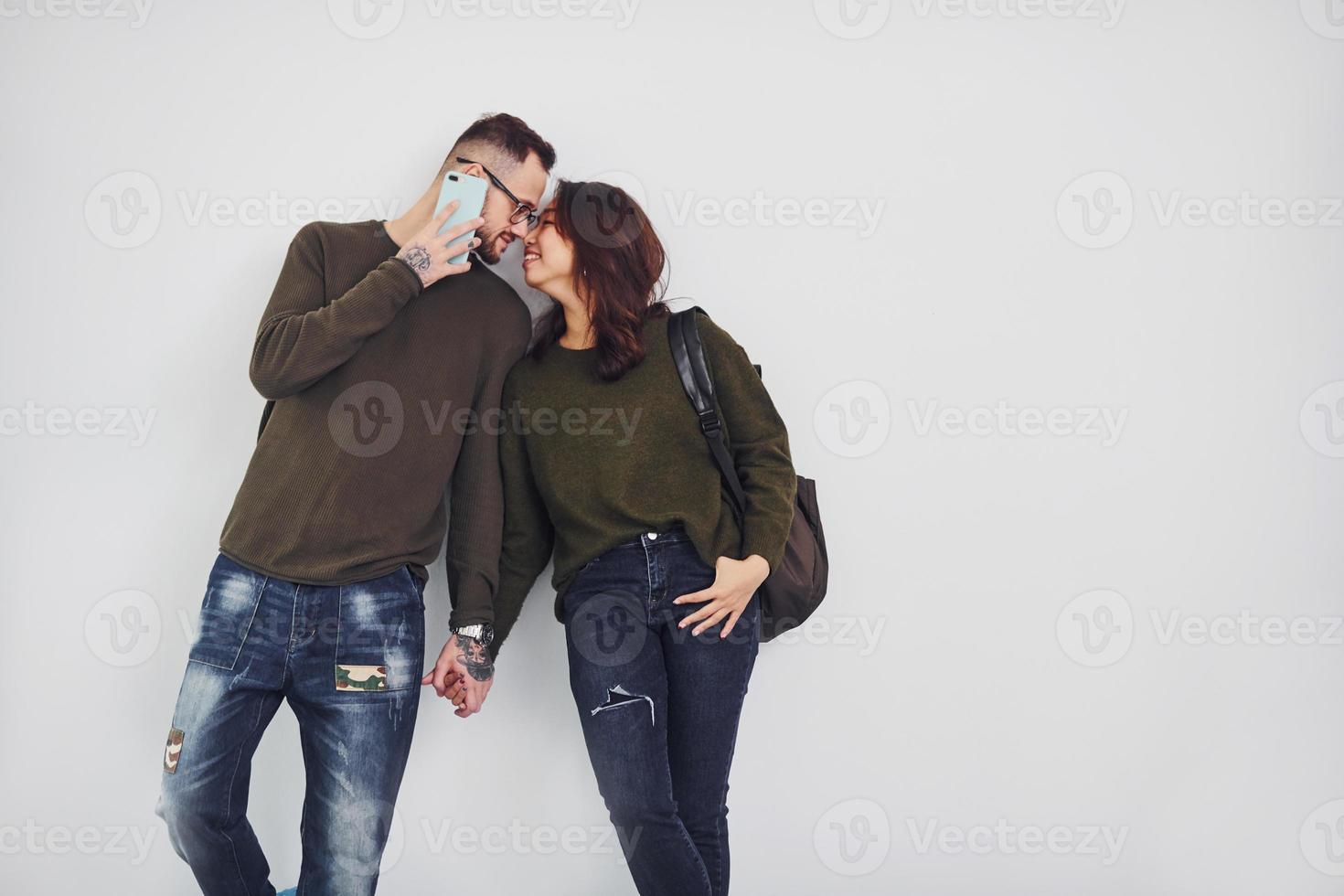 Cheerful multi ethnic couple with backpack and phone standing together indoors in the studio against white background photo