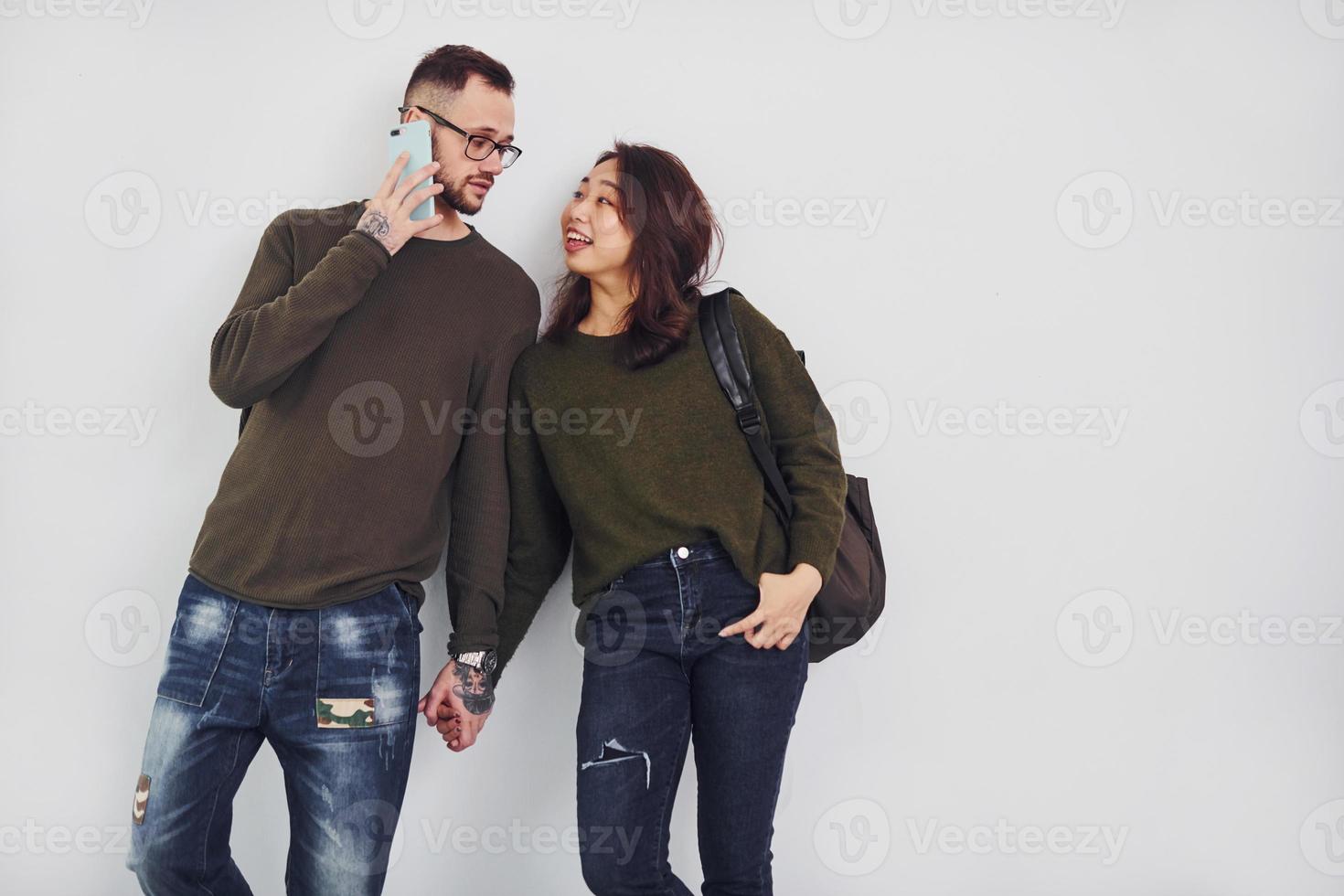 Cheerful multi ethnic couple with backpack and phone standing together indoors in the studio against white background photo