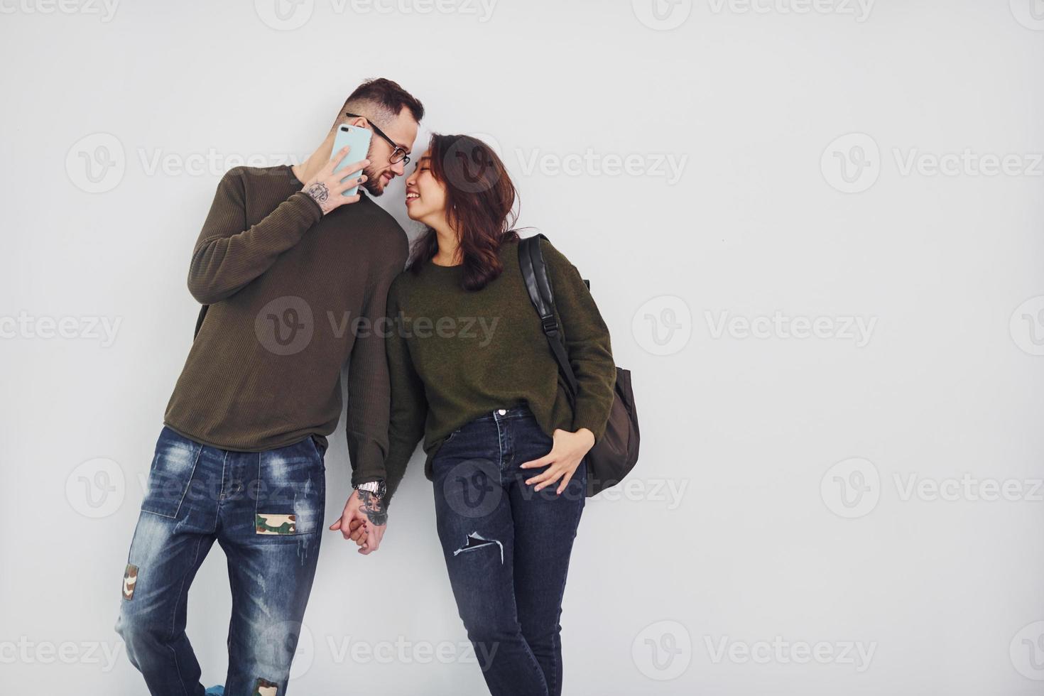 alegre pareja multiétnica con mochila y teléfono de pie juntos en el interior del estudio con fondo blanco foto