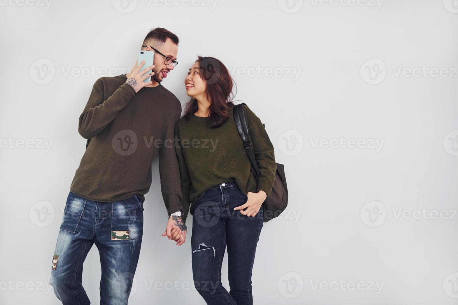 Cheerful multi ethnic couple with backpack and phone standing together indoors in the studio against white background photo