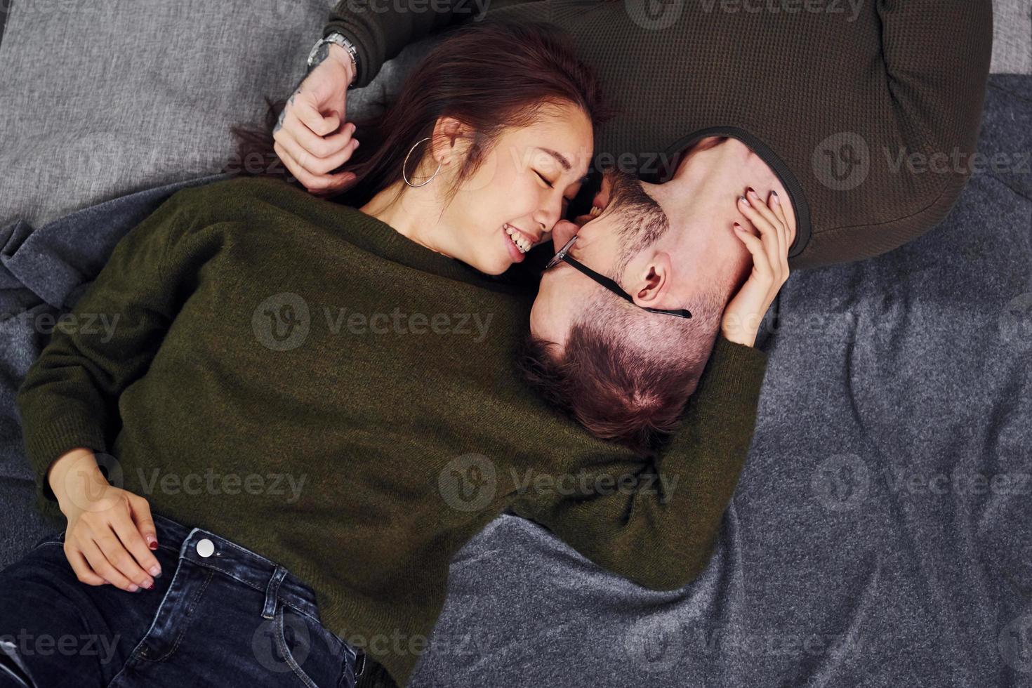 Happy multi ethnic couple in casual clothes lying down together indoors in the studio. Caucasian guy with asian girlfriend photo