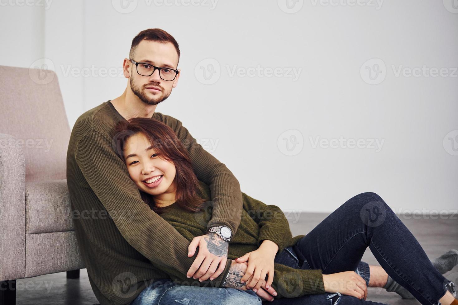 Happy multi ethnic couple in casual clothes sitting together indoors in the studio. Caucasian guy with asian girlfriend photo