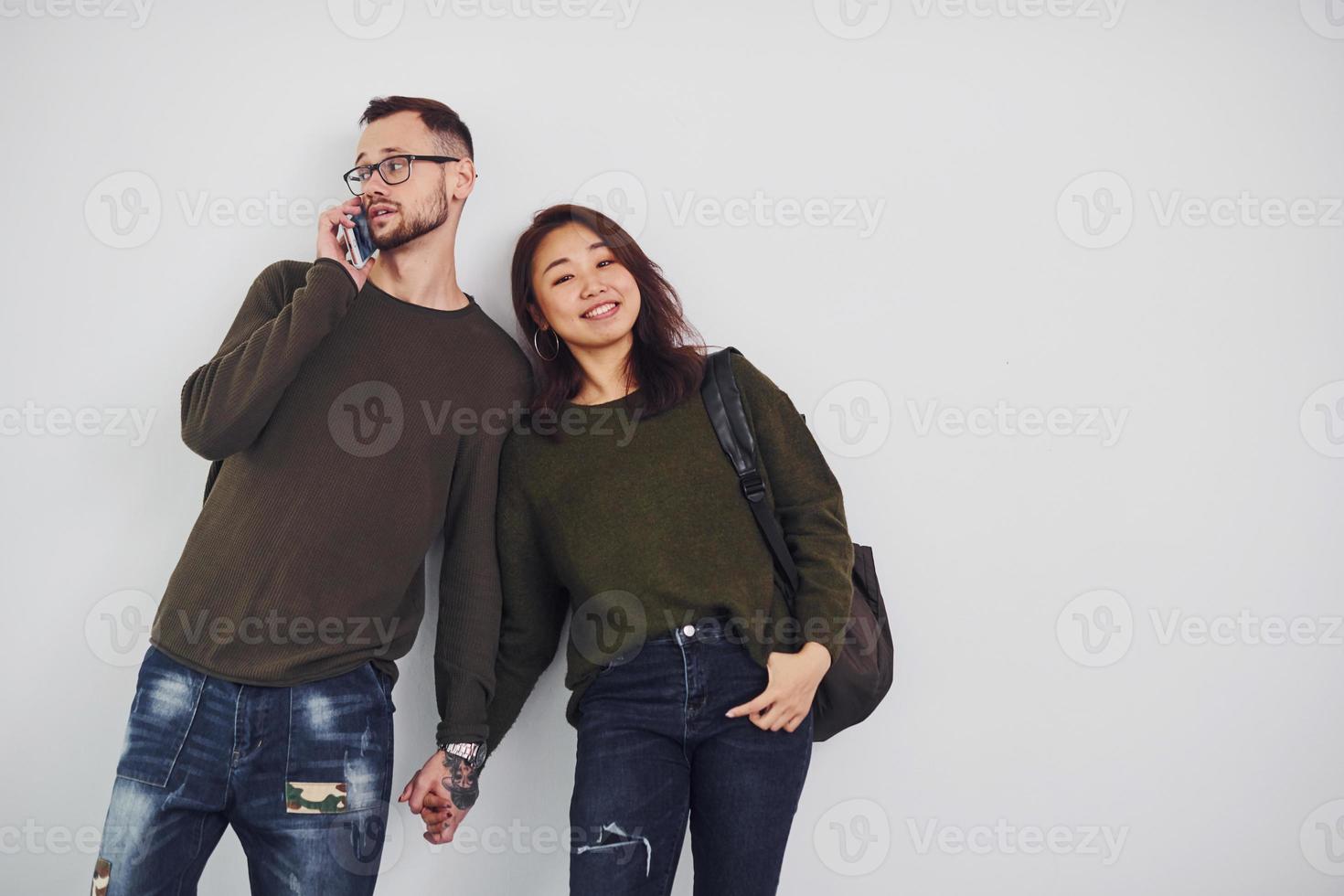 Cheerful multi ethnic couple with backpack and phone standing together indoors in the studio against white background photo