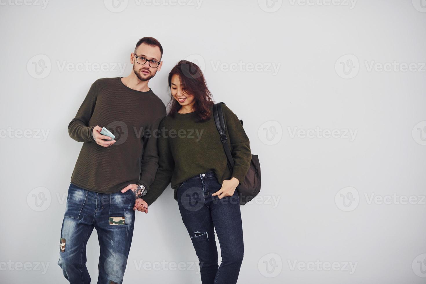 Cheerful multi ethnic couple with backpack and phone standing together indoors in the studio against white background photo
