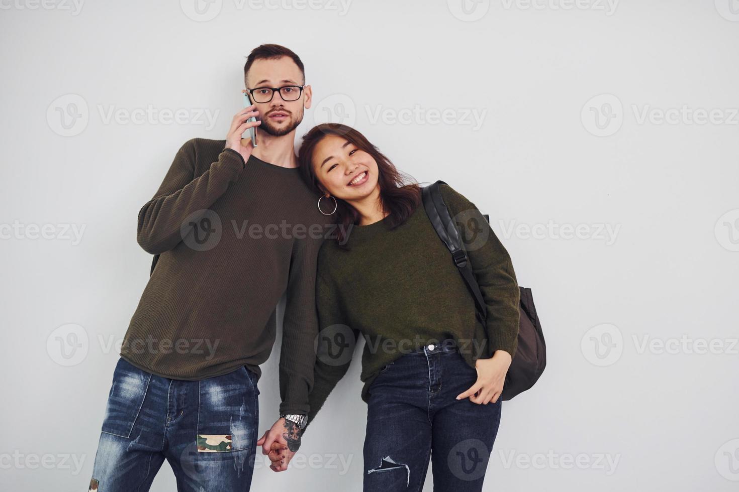 Cheerful multi ethnic couple with backpack and phone standing together indoors in the studio against white background photo