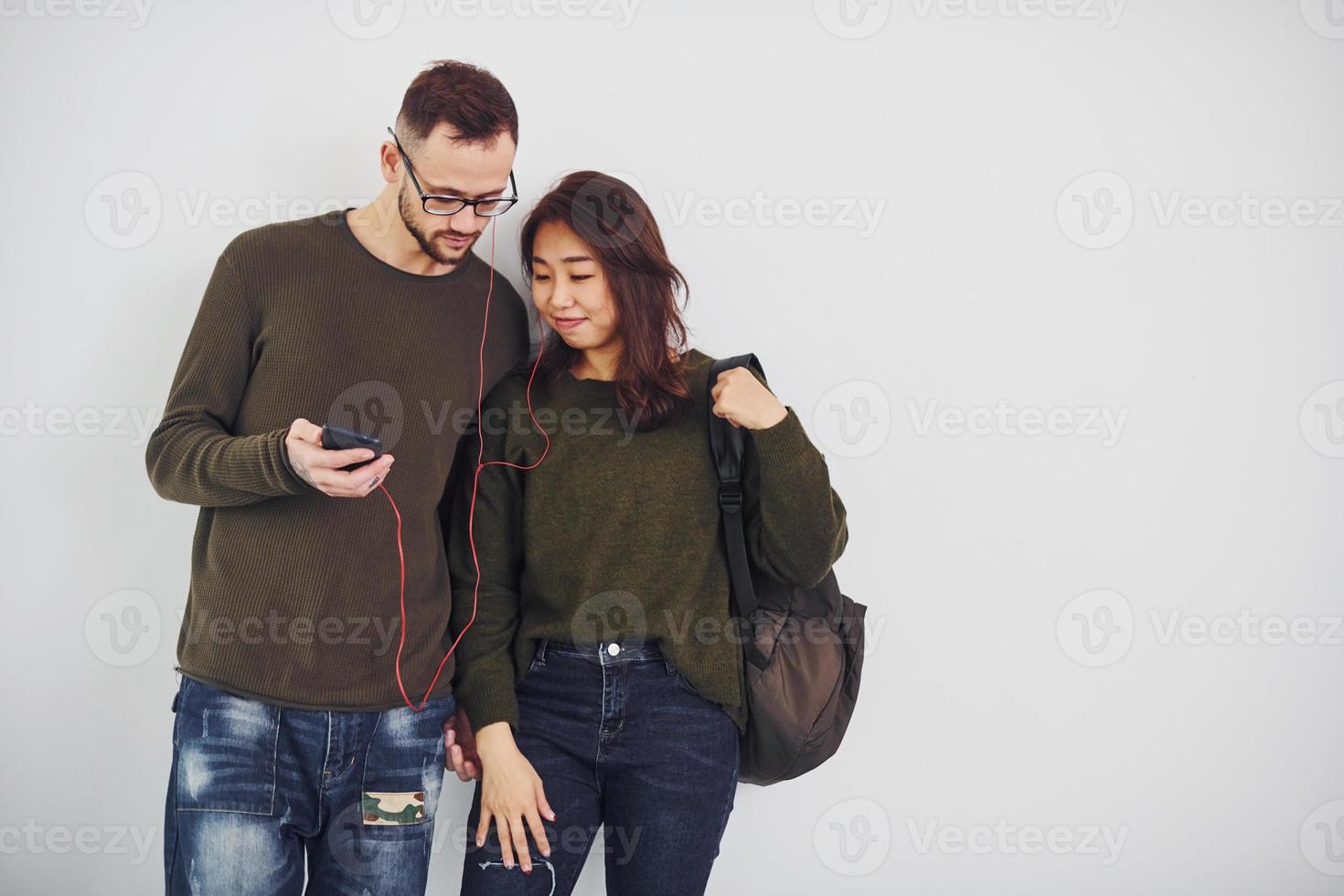 Cheerful multi ethnic couple with backpack and phone standing together indoors in the studio against white background photo