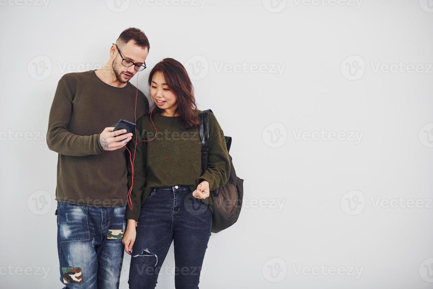 Cheerful multi ethnic couple with backpack and phone standing together indoors in the studio against white background photo