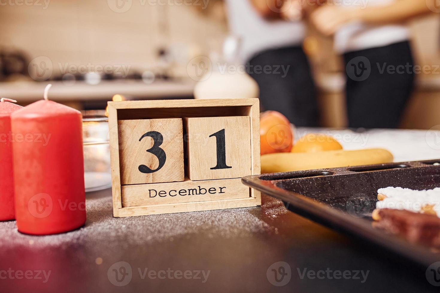 Wooden calendar with 31 december writen on it. Candles and food on the table. People at background photo
