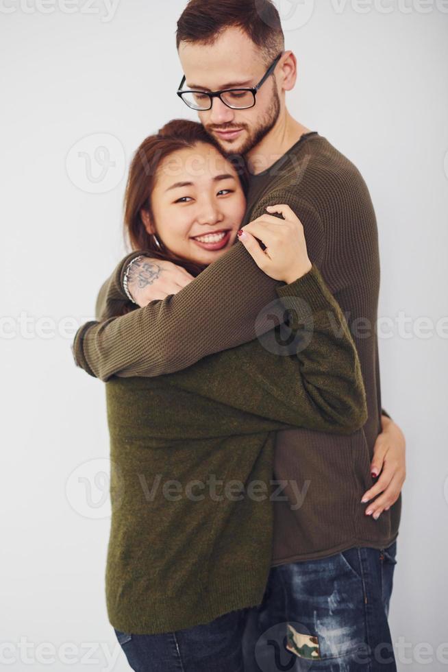 feliz pareja multiétnica con ropa informal abrazándose unos a otros en el interior del estudio. chico caucásico con novia asiática foto