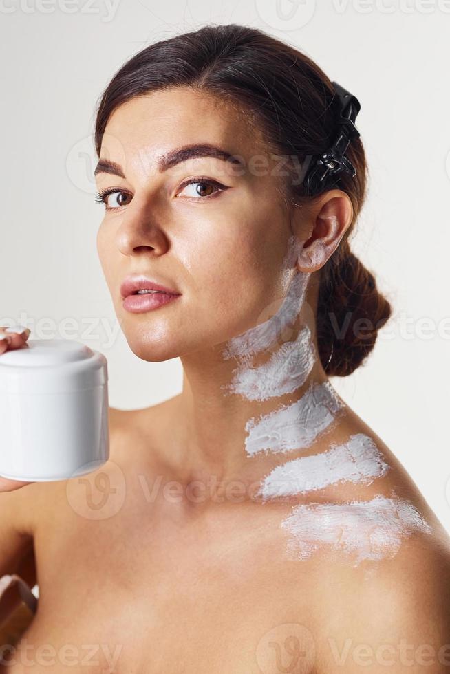 Naked woman with paint on neck is in the studio against white background with jar photo