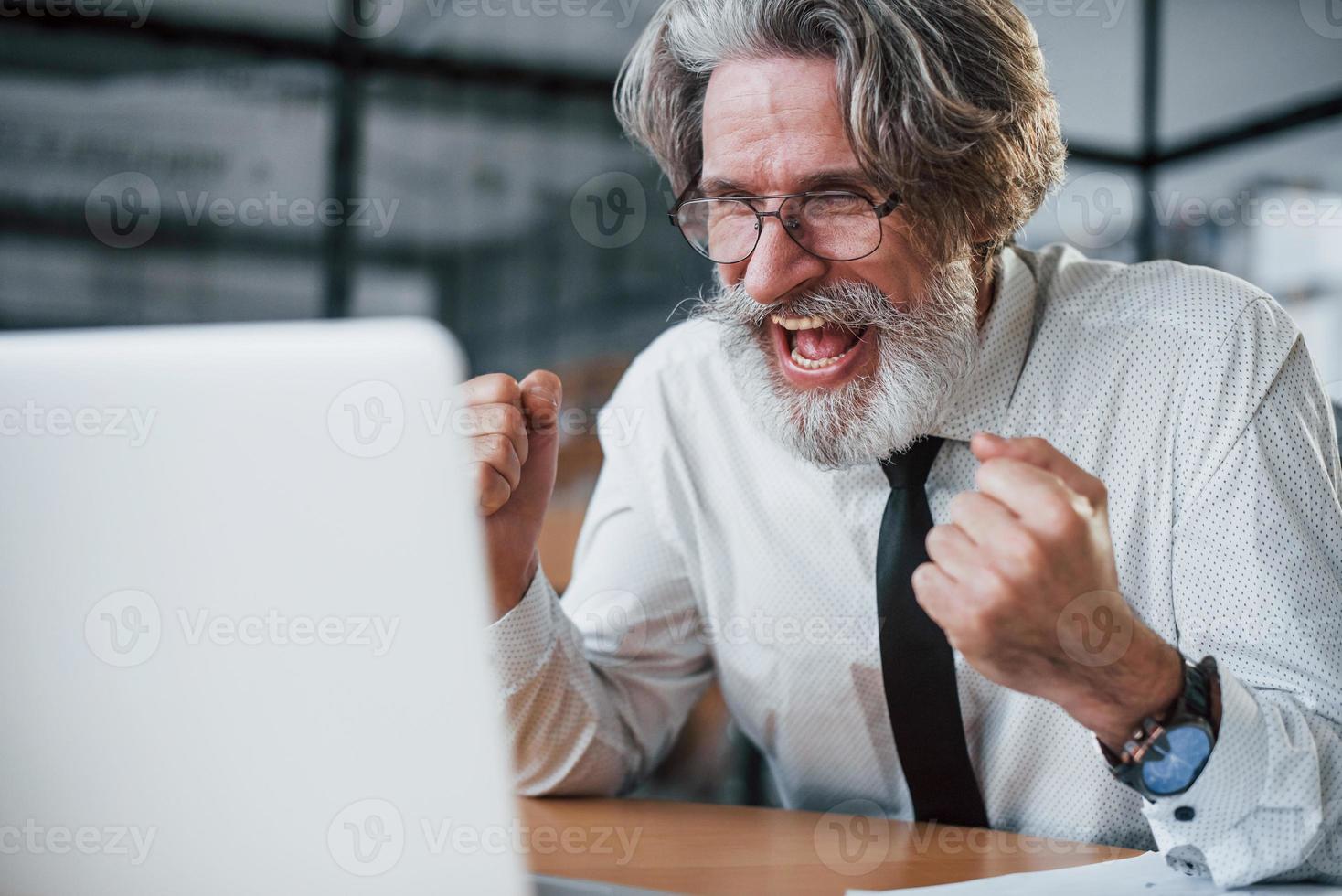 Expressive mature businessman with grey hair and beard in formal clothes is in the office against laptop photo