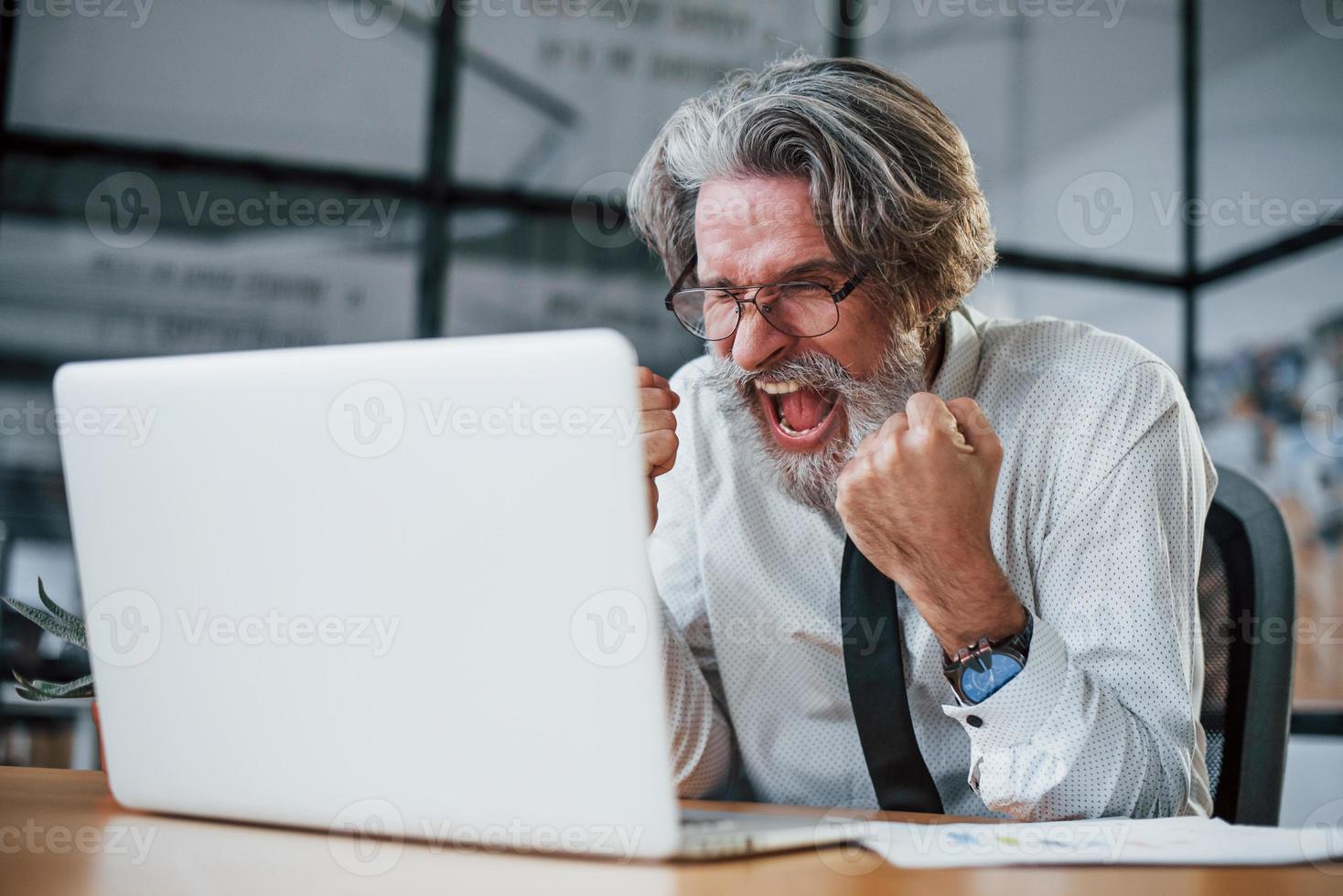 Expressive mature businessman with grey hair and beard in formal clothes is in the office against laptop photo